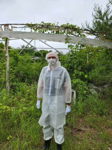 Photograph of a man wearing a full, white protective suit and mask in an overgrown yard, in front of a ruined structure.