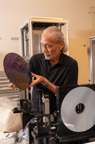 A scientist looks at a circular silicon wafer etched with circuitry, with his face reflected on the surface of the wafer. In the foreground, a glimpse of the hardware for in-line EUV lithography is visible.