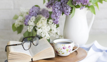 open book on a table, with eyeglasses on top. a cup of coffee on a try and a vase of purple and white flowers.