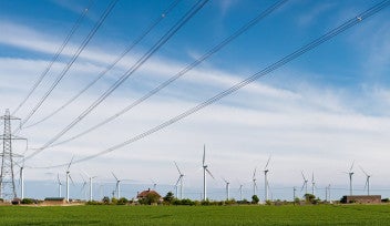Panoramic photograph of a tall transmission line, with a wind farm in the background against a blue sky.