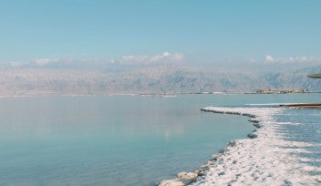 Salt crystals are exposed along the edge of a large, serene body of water. The water reaches some far-off, cloud-capped mountains in the background.