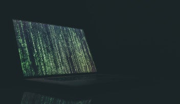A laptop screen is illuminated against a dark background. The screen is lit up with vertical lines of green code.