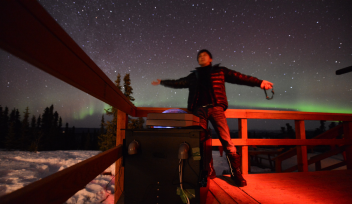 Dr Ryuho Kataoka stands arms spread wide under a bright starry-sky featuring a green aurora. He is next to a fence and some scientific equipment in a snowy tree-lined field. 