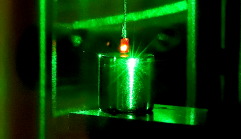 Photograph with a small square diamond glowing red as a result of being illuminated with a green laser, suspended above a smooth cylindrical magnet.