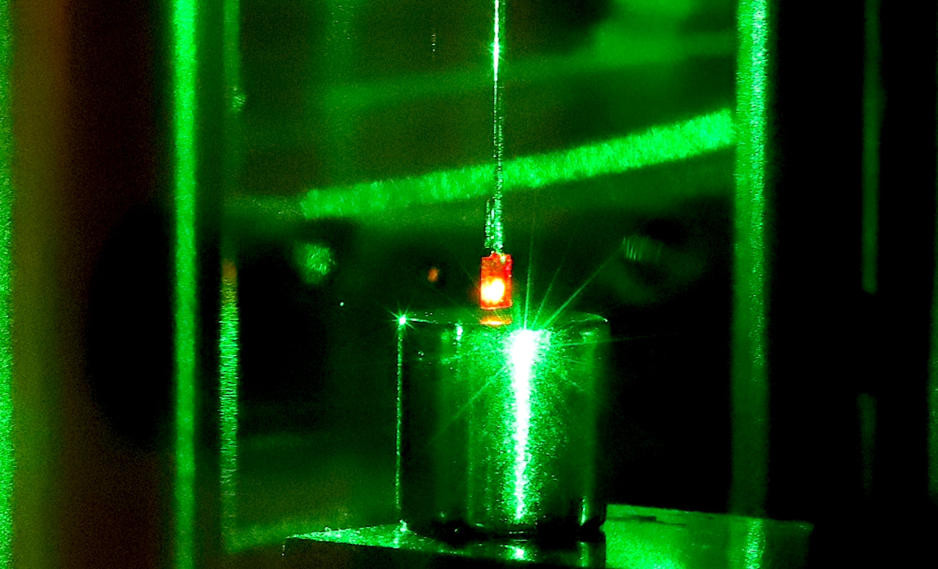 Photograph with a small square diamond glowing red as a result of being illuminated with a green laser, suspended above a smooth cylinder.