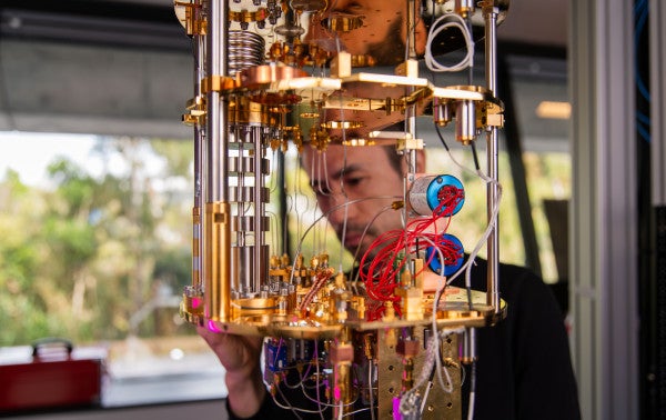 A beautiful gold machine with lots of wires in the foreground, at which a man in the background stares intently. 